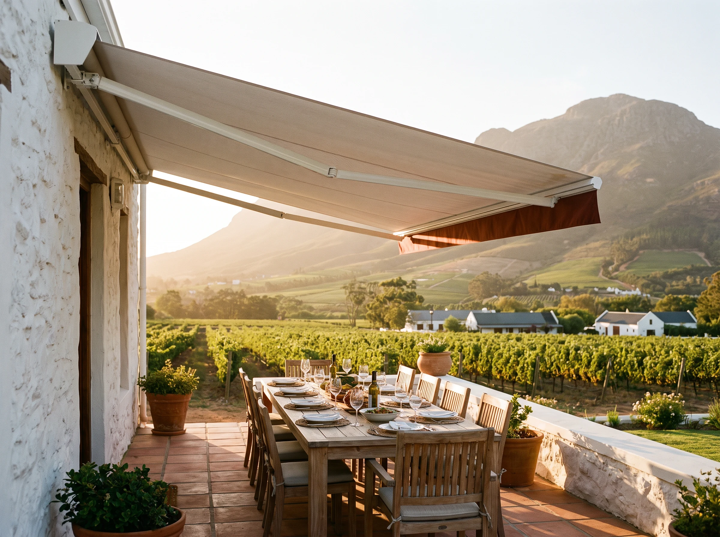 Retractable folding-arm awning shading an outdoor dining table on a farmhouse stoep near Klapmuts, vineyard rows and Simonsberg mountain in the background