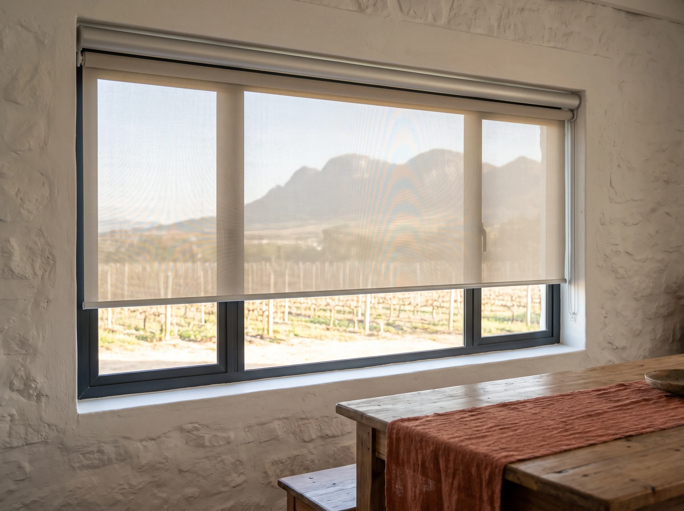 Sunscreen roller blind over a wide window in a Klapmuts farmhouse dining room, vineyard rows and Simonsberg mountain visible through the mesh