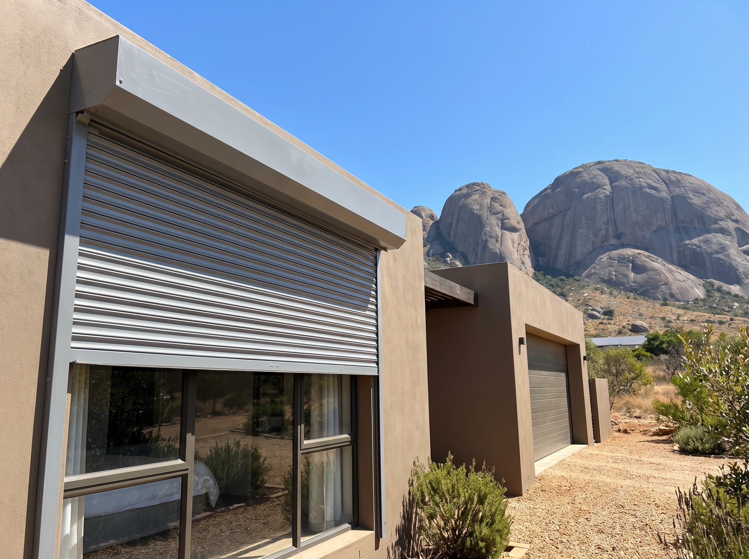 External roller shutter with rigid aluminium slats partially lowered over a west-facing window on a Paarl home, Paarl Rock's granite domes visible behind